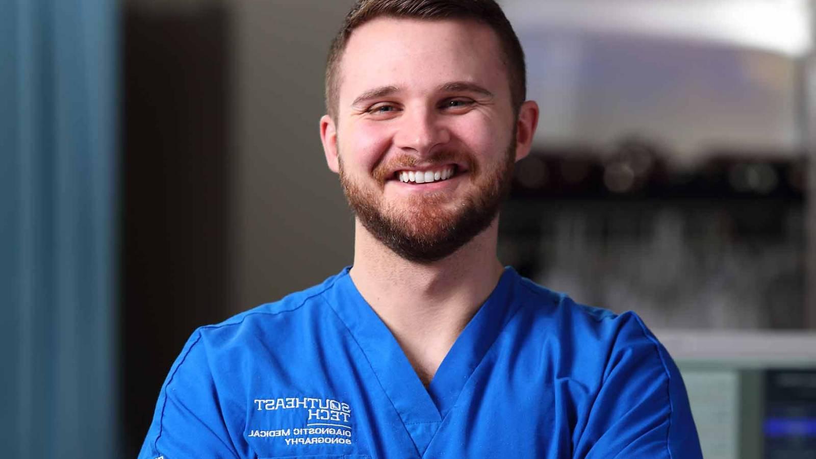 Male sonography student wearing blue scubs in exam room, smiling